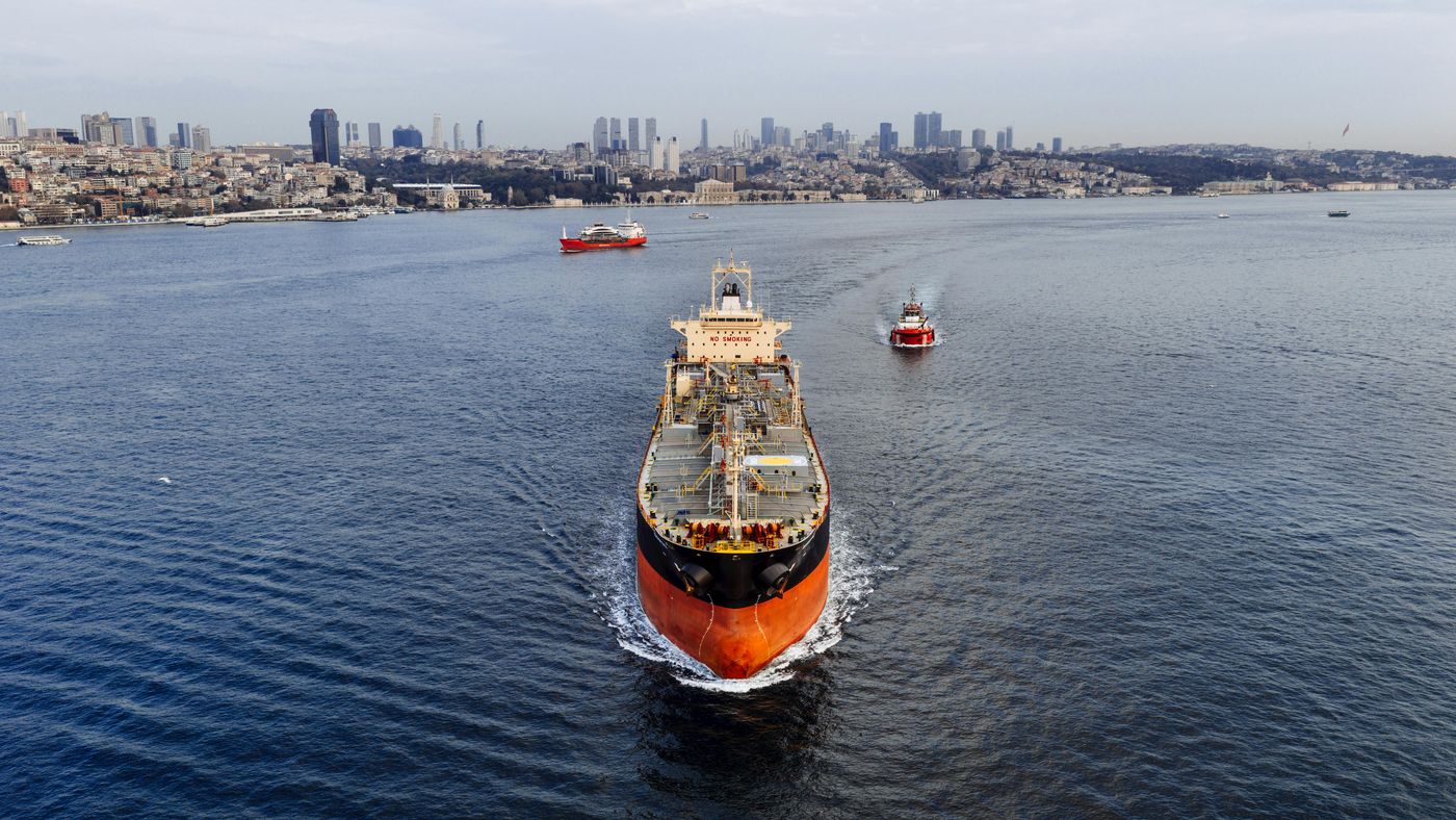 A loaded tanker proceeds along a busy waterway with a pilot launch in attendance and a coastal city skyline beyond.